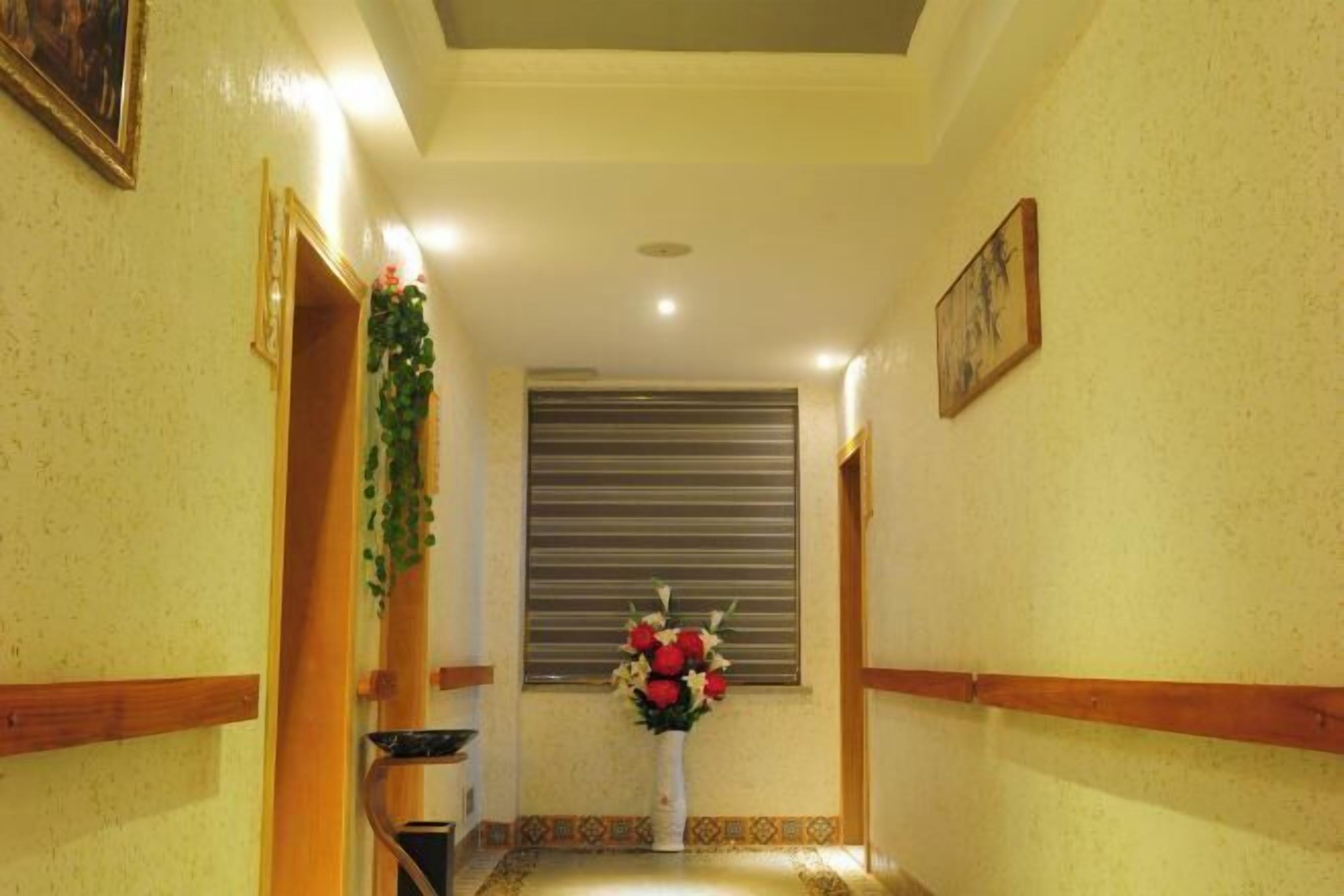 A hallway with a decorative vase of flowers, a window with blinds, and a framed picture on the wall.