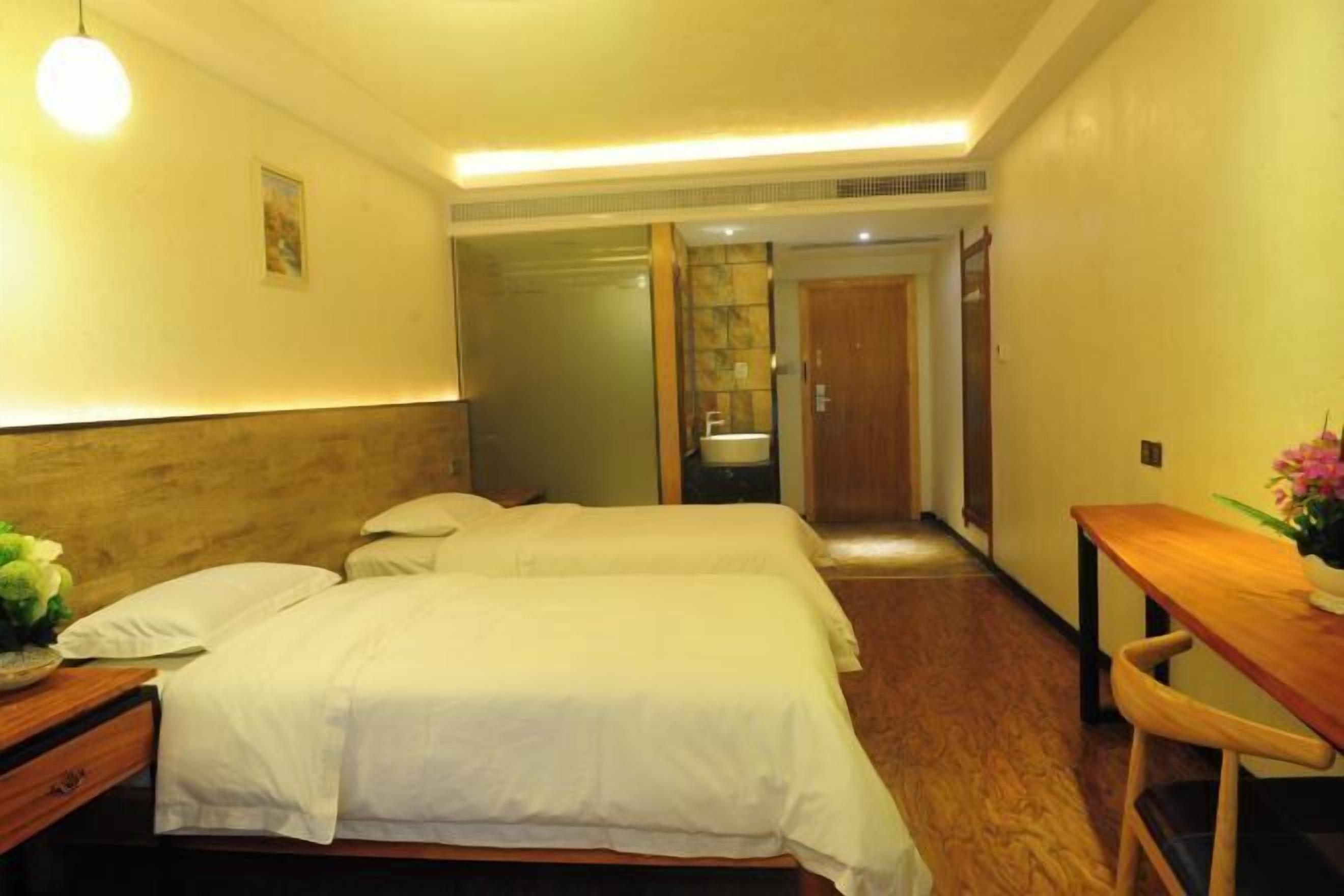 Ein Hotelzimmer mit zwei Betten, einem Holztisch, einem Stuhl, einem kleinen Tisch mit einer Vase und einem durchgegangenen Tür erkennbaren Badezimmer.