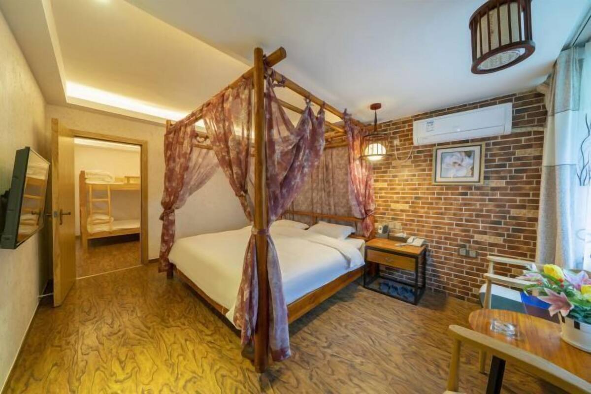 A room with a four-poster bed, a wooden floor, a brick wall, an air conditioning unit, and a small table with a vase of flowers.