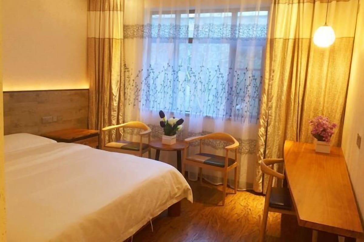 A hotel room with a bed, two chairs, a small table, a vase of flowers, and a window with curtains.