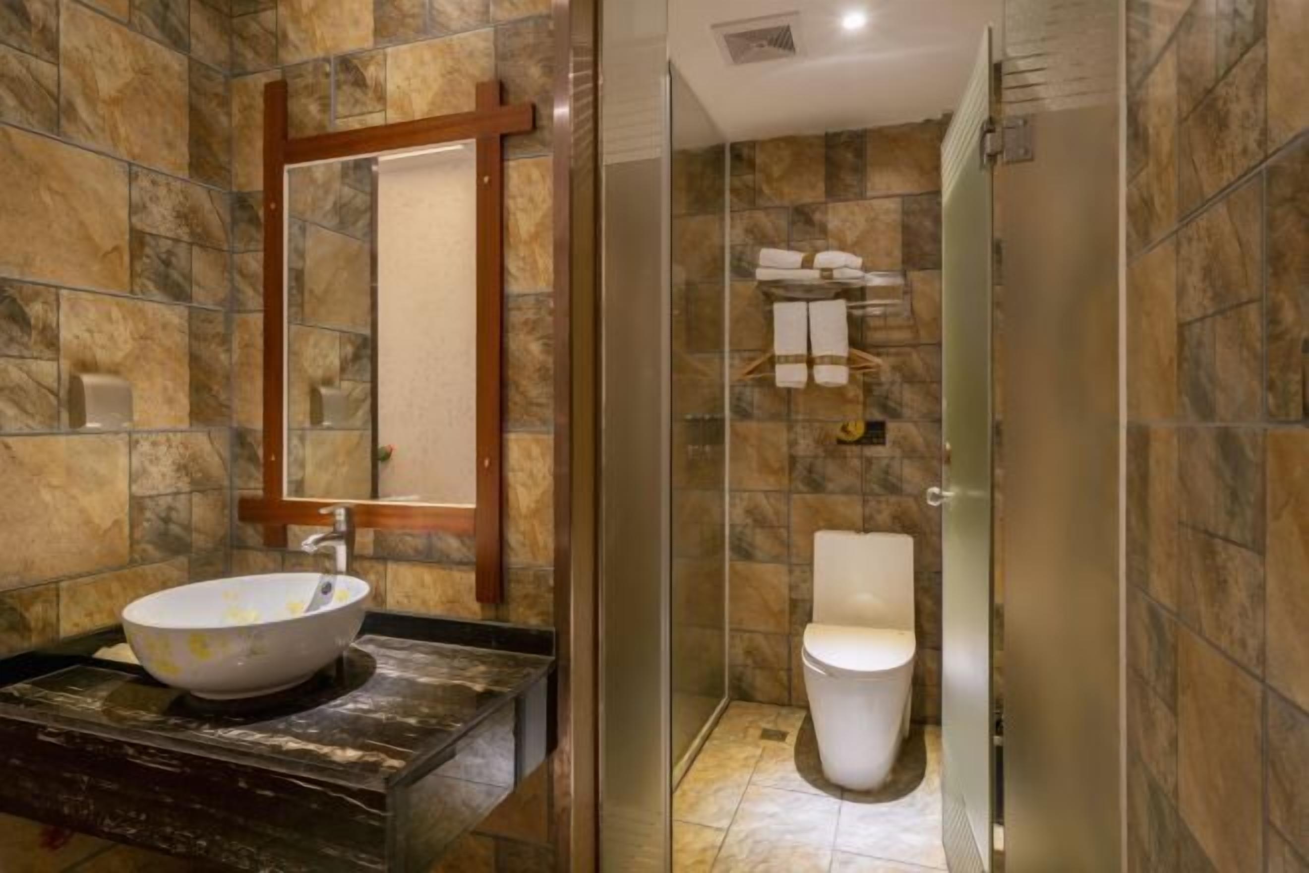 Un baño con pared de baldosas de piedra, un espejo con marco de madera, un lavabo blanco y una zona de ducha con mampara de vidrio.
