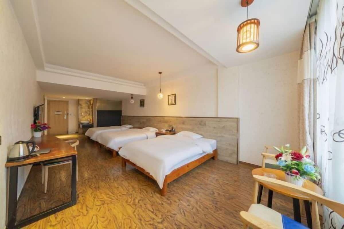 A hotel room with three beds, a wooden desk, a small table with flowers, and a wall-mounted TV.