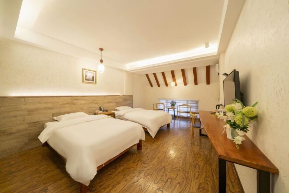 A hotel room with two beds, a wooden floor, a desk with a vase of flowers, and a large window.