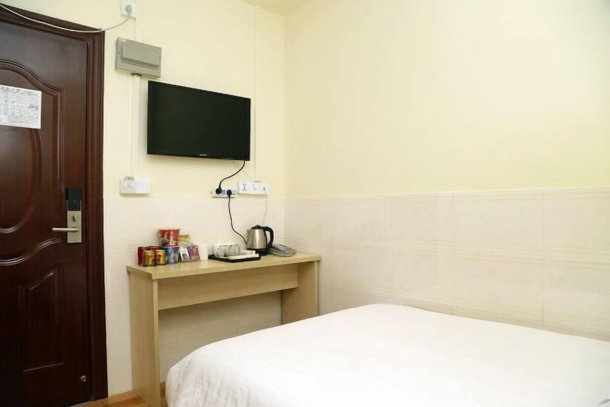A hotel room with a bed, a television mounted on the wall, a desk with a kettle and a phone, and a wooden door.
