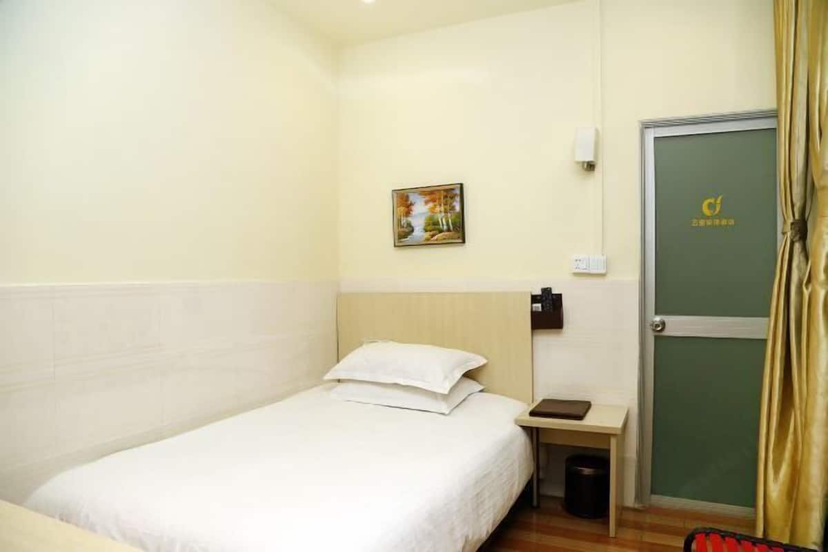 A hotel room with a bed, a small bedside table, a trash bin, a painting on the wall, and a door with a logo.