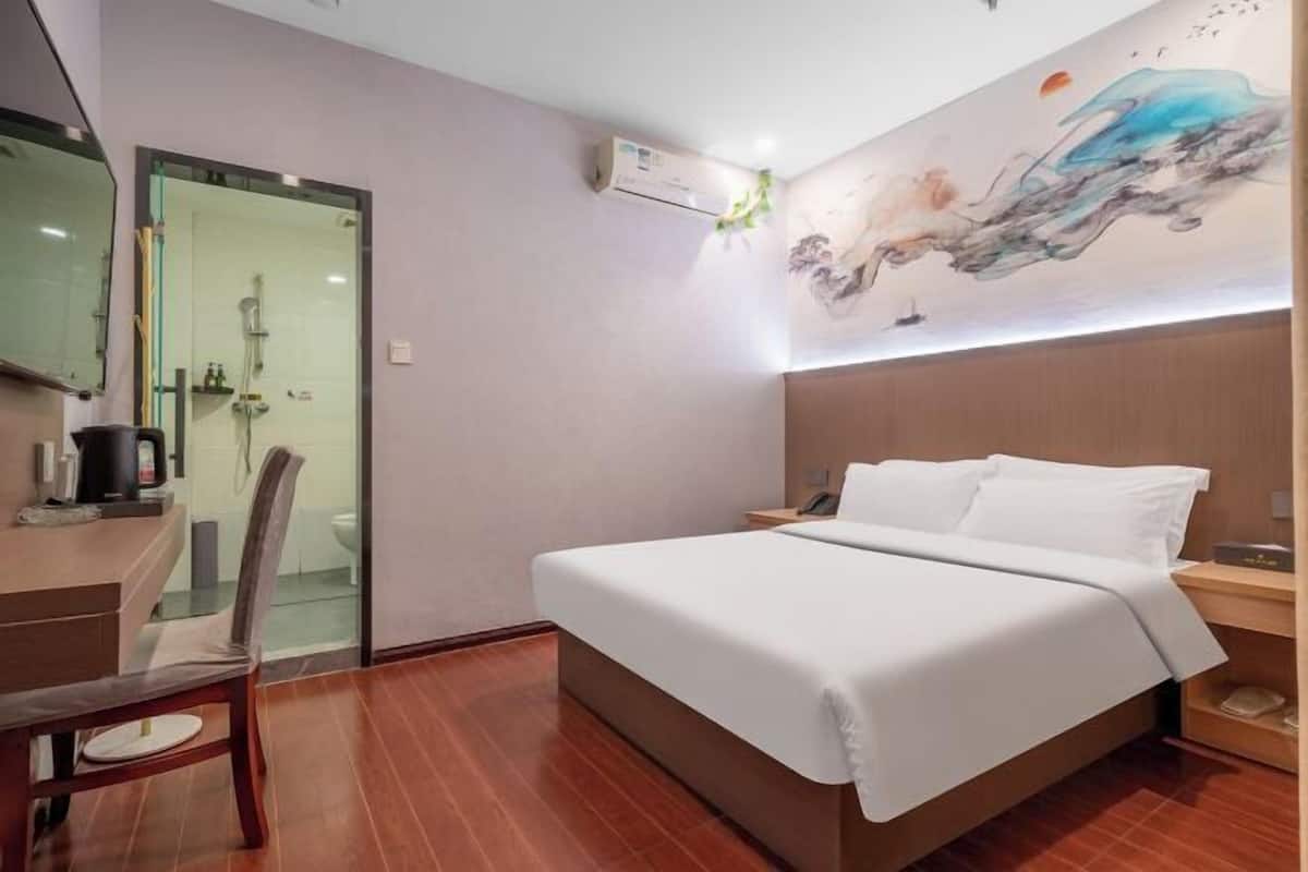 A hotel room with a bed, a desk, a chair, a shower area, and a wall with a painting.