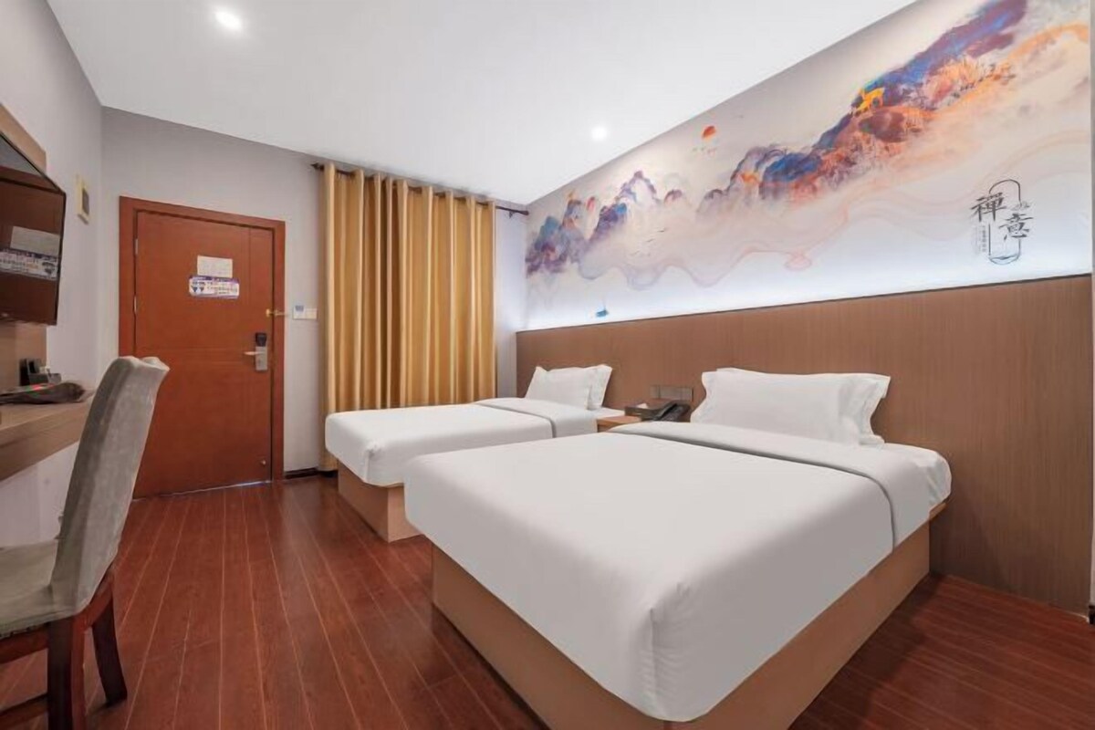 A hotel room with two beds, a desk, a chair, a door, and a wall with a colorful abstract painting.