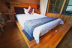 A wooden-framed bed with a patterned bedspread and pillows, a bedside table with a lamp, and a window with a view of greenery. - Padma Chumig Garden Hotel (Lhasa)