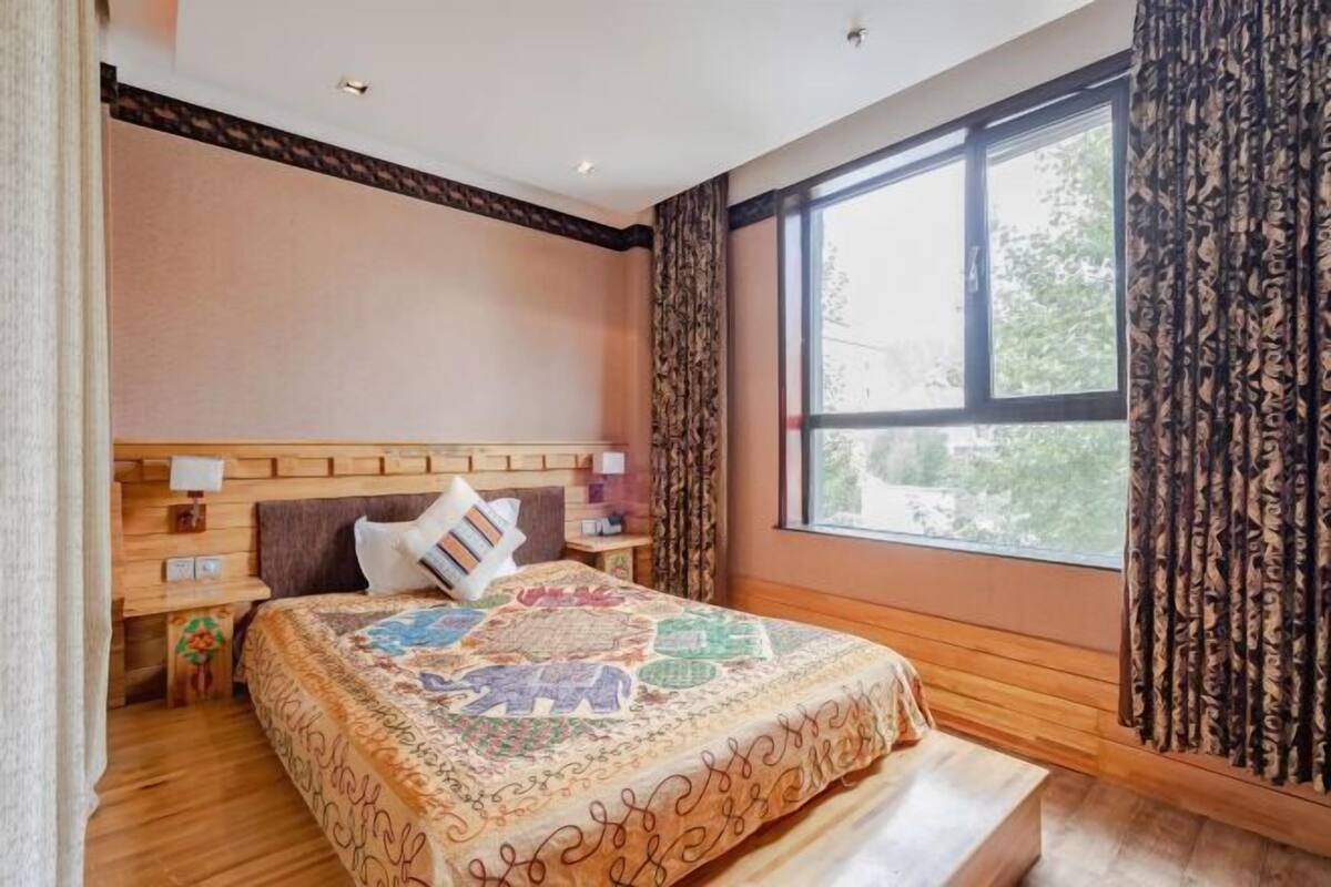 Een slaapkamer met een houten hoofdbord, een groot raam met gordijnen, een bed met een bedovertrek met patroon en een nachtkastje met een lamp.