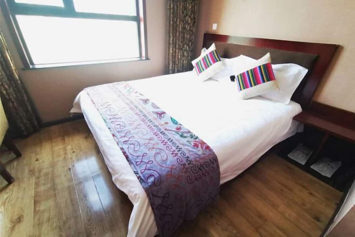 Een hotelkamer met een houten vloer, een bed met een witte dekbedovertrek en kleurrijke kussens, een raam met gordijnen, en een houten hoofdeinde.
