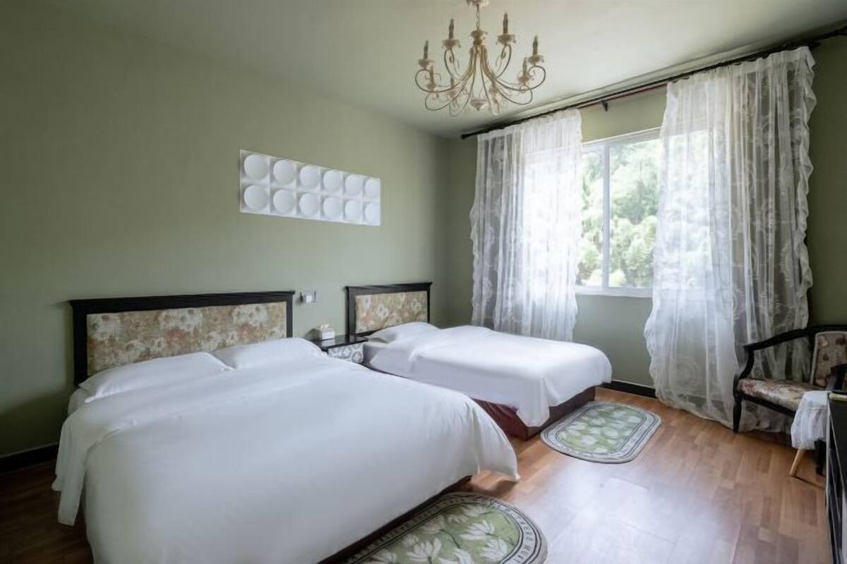 Quarto com duas camas, um candelabro, janela com cortinas leves e uma cadeira.