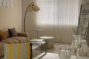 Un salon avec un canapé, une table basse, un lampadaire et une fenêtre avec des stores.
