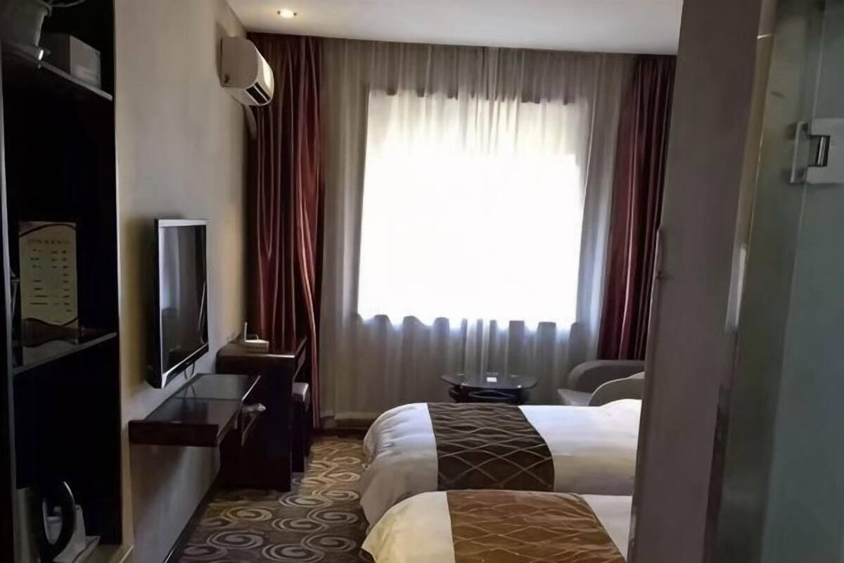 Ein Hotelzimmer mit zwei Betten, einem Schreibtisch, einem Flachbildfernseher, einem Sessel und einem Vorhangfenster.