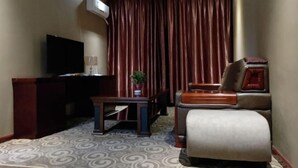 Ein Hotelzimmer mit Flachbildfernseher, Schreibtisch mit Lampe, Sessel, kleinem Tisch mit Pflanze und einem gemusterten Teppich.