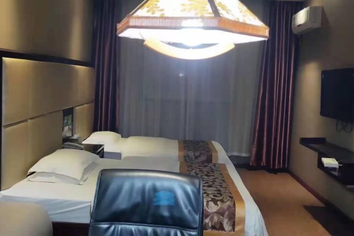 Ein Hotelzimmer mit zwei Betten, einer großen Lüster, einem Flachbildfernseher und einem mit Vorhängen versehenen Fenster.