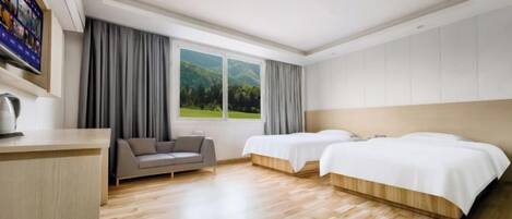 A hotel room with a wooden floor, two beds, a sofa, a TV, and a window with a view of greenery.
