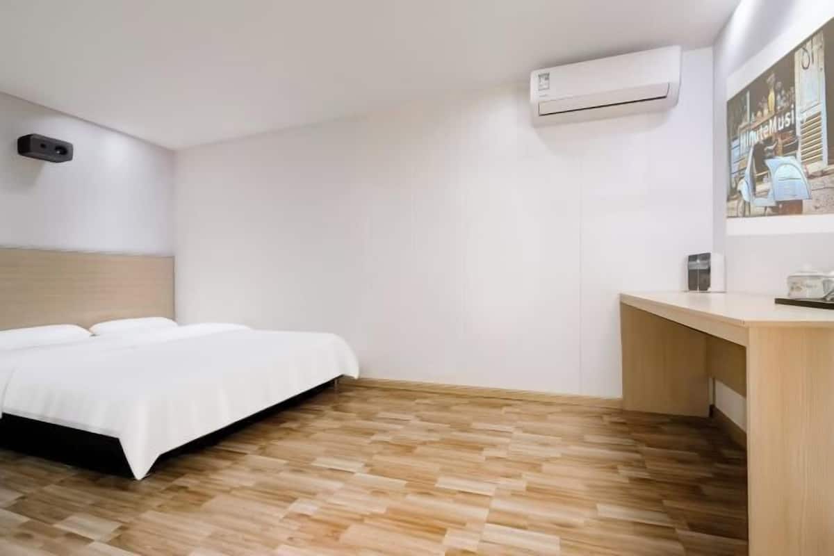 A hotel room with a bed, wooden flooring, a desk, and an air conditioning unit.
