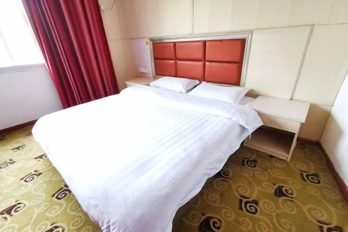 A hotel room with a bed, a nightstand, and a window with red curtains.