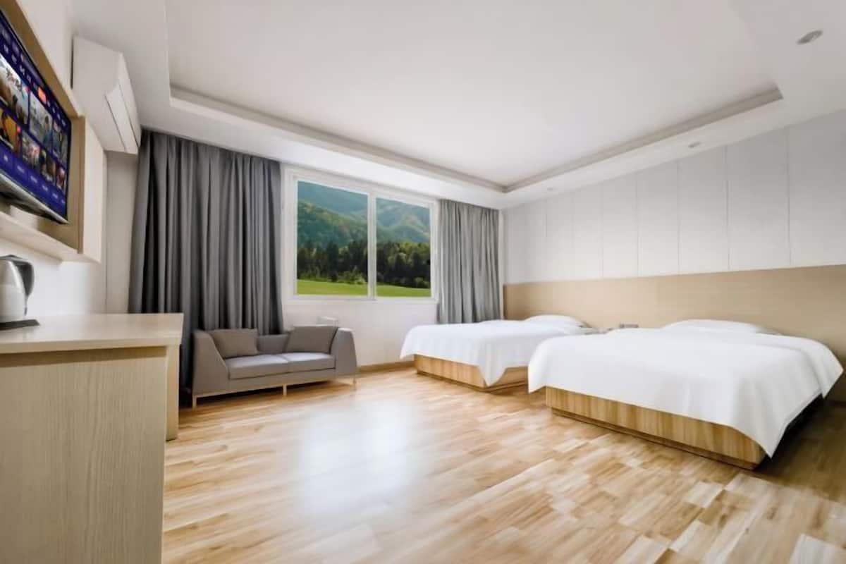 A hotel room with a wooden floor, two beds, a sofa, a TV, and a window with a view of greenery.