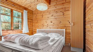 Ein Schlafzimmer einer Holzhütte mit einem Bett, weißer Bettwäsche, einem Fenster mit Vorhängen und einer Pendelleuchte.