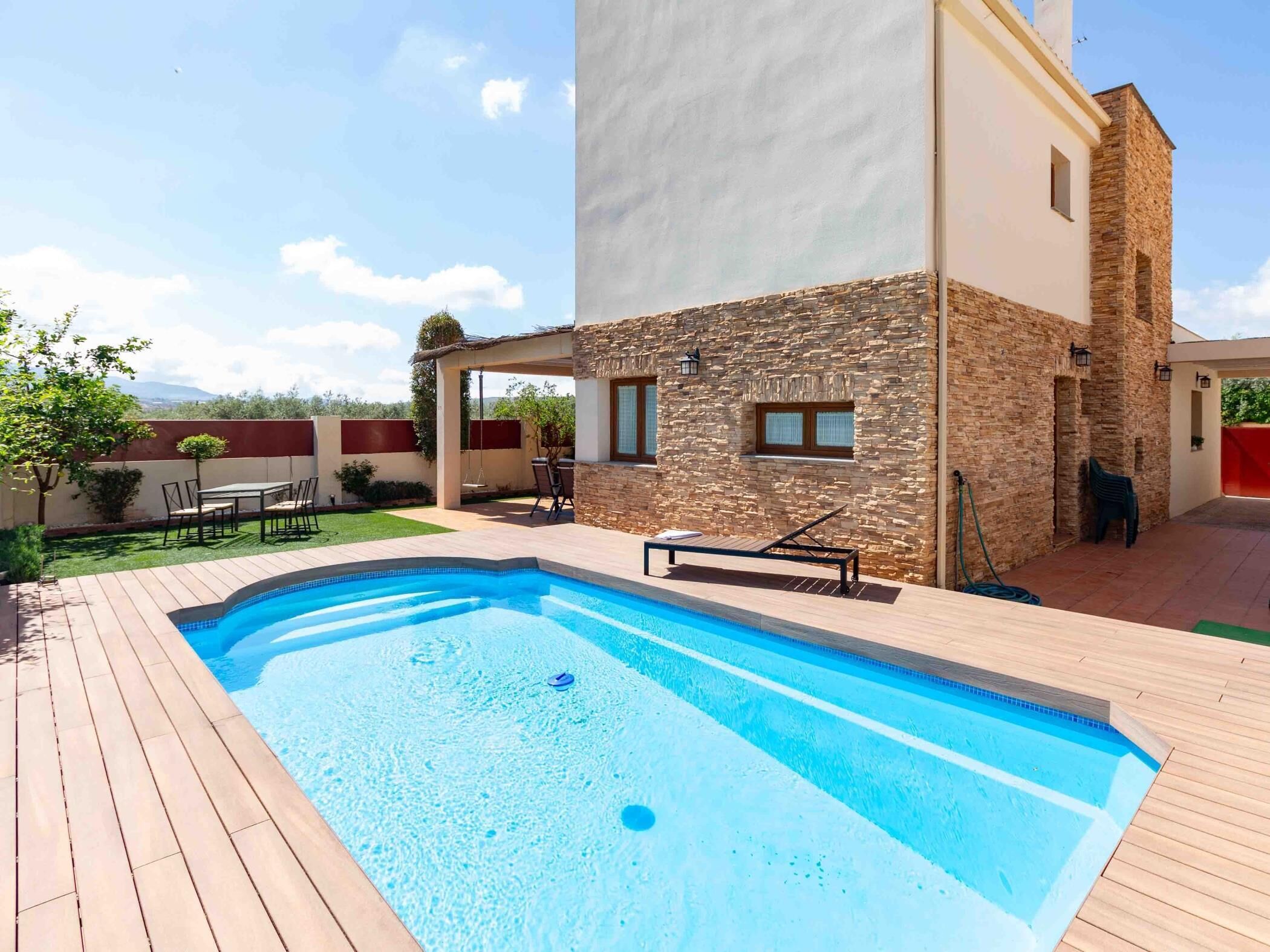 A swimming pool with a wooden deck, a stone wall, and a covered seating area.