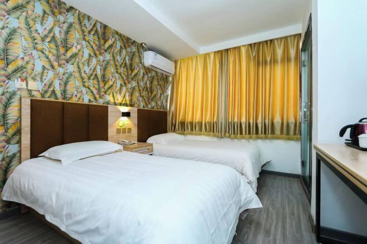 A hotel room with two beds, a wooden headboard, a nightstand with a lamp, a window with yellow curtains, and an air conditioning unit.