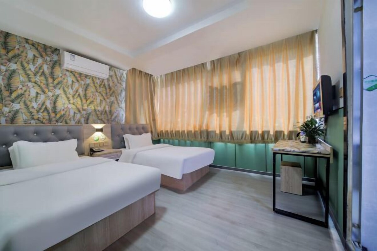 A hotel room with two beds, a bedside table with a lamp, a TV on the wall, and a window with curtains.