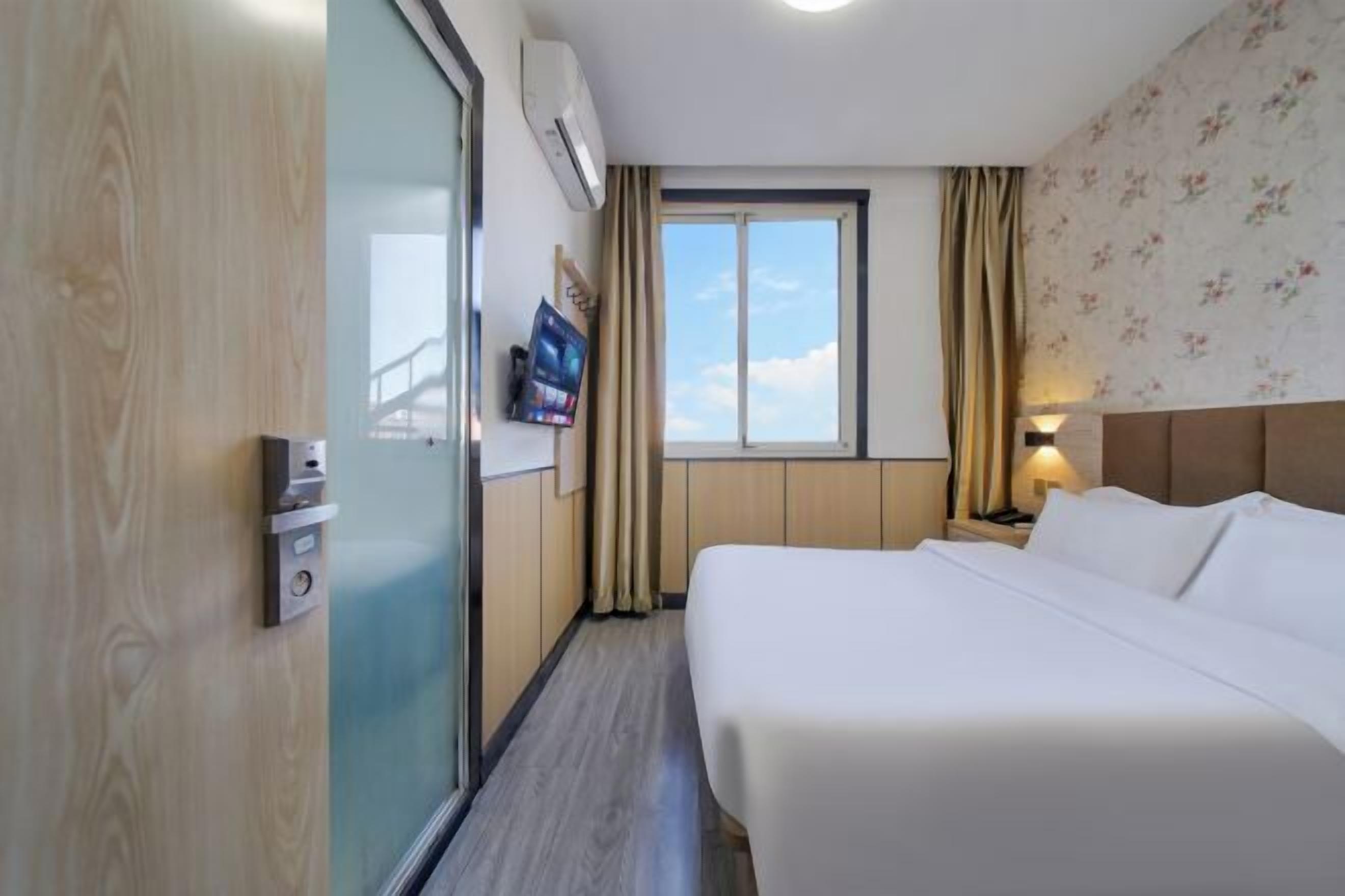 Una habitación de hotel con cama, televisión montada en la pared, ventana con cortinas y puerta de madera.