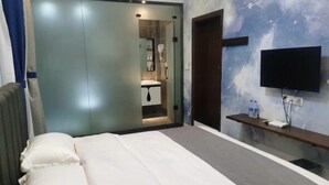 A bedroom with a bed, a TV mounted on the wall, a small shelf with items, and a door leading to a bathroom. - Lujiang Business and Trade City Hotel (Rongjiang)