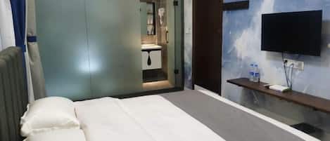 Un dormitorio con cama, televisor montado en la pared, un estante con objetos y una puerta que da a un baño.