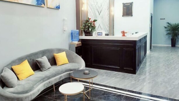 A modern reception area with a grey sofa, a small round table, a marble floor, and a reception desk with a plant. - Lujiang Business and Trade City Hotel (Rongjiang)