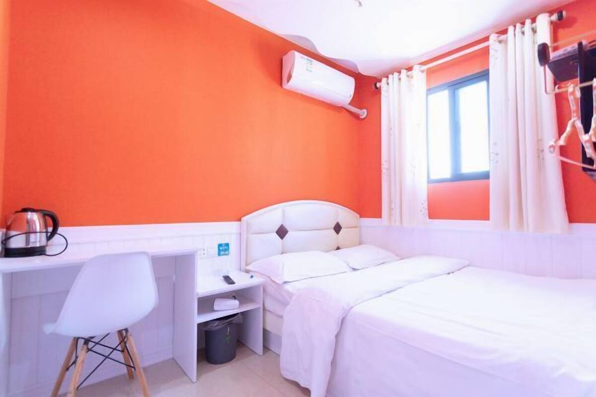 A hotel room with a white bed, a desk with a chair, a radiator, and a window with curtains.
