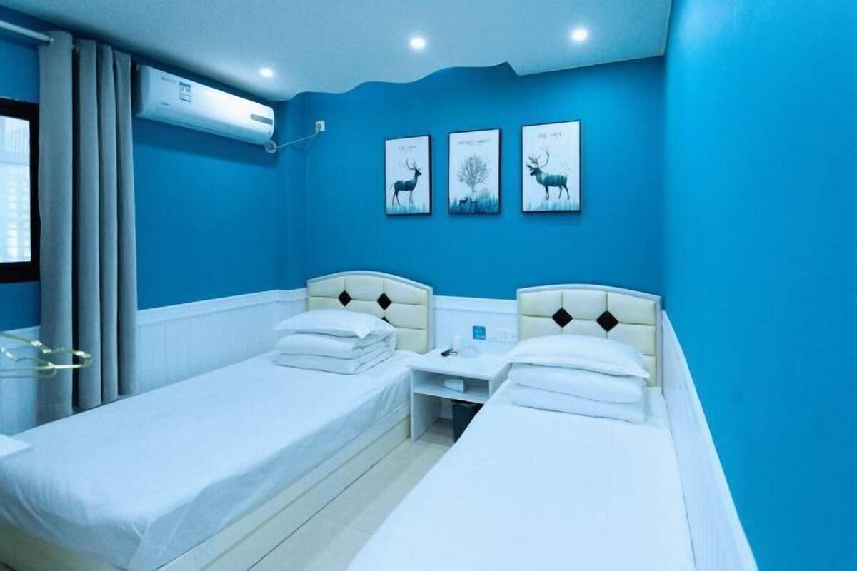 A hotel room with two single beds, a bedside table, a wall-mounted air conditioner, and three framed pictures on the wall.