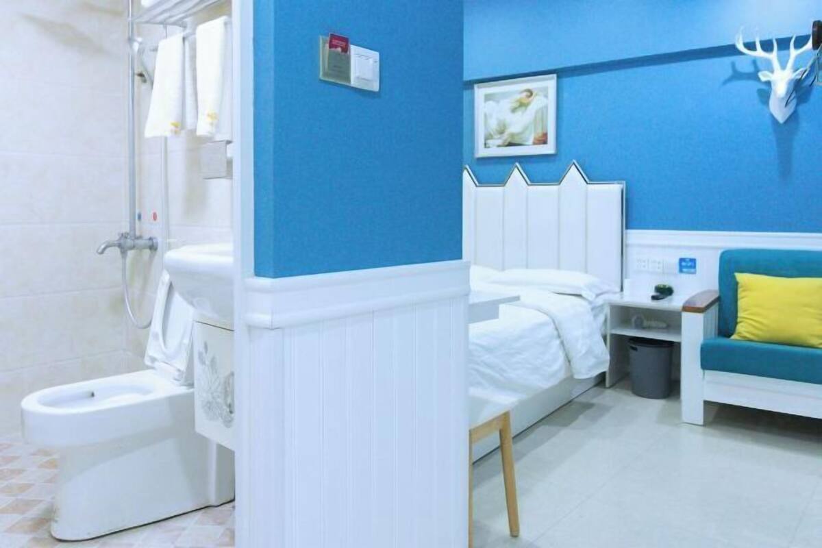 A bathroom with a toilet, bidet, and shower. There is a bed with a white headboard and a blue and yellow sofa.