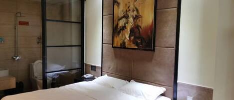 A hotel room with a bed, a large painting on the wall, a bathroom with a glass shower enclosure, and a small bedside table.