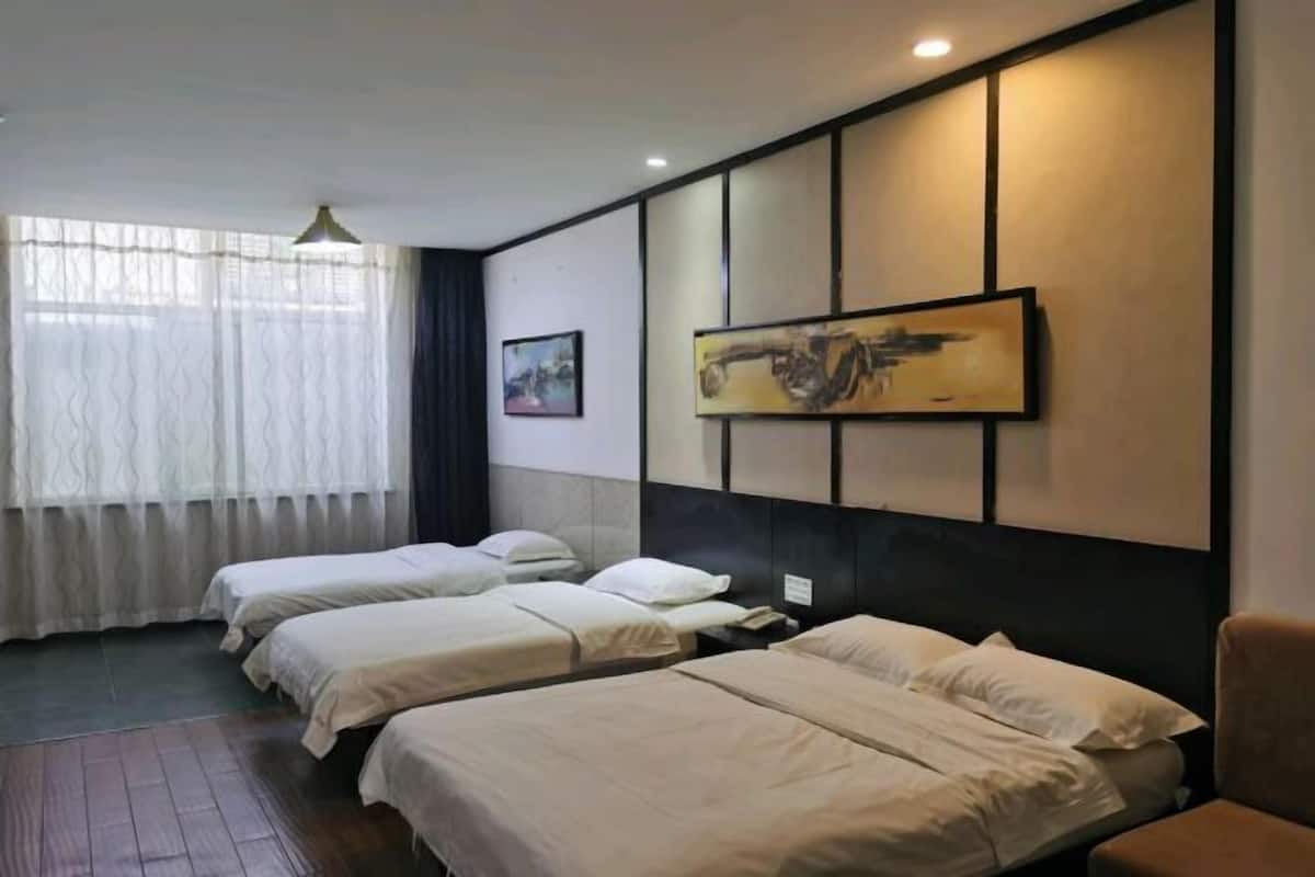 A hotel room with two beds, a large window with sheer curtains, a painting on the wall, and a ceiling light.