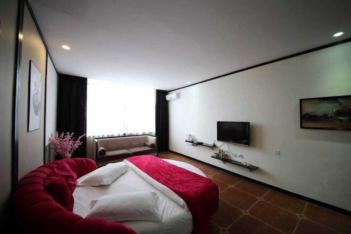 A modern hotel room with a large bed, a sofa, a TV mounted on the wall, and a window providing natural light.