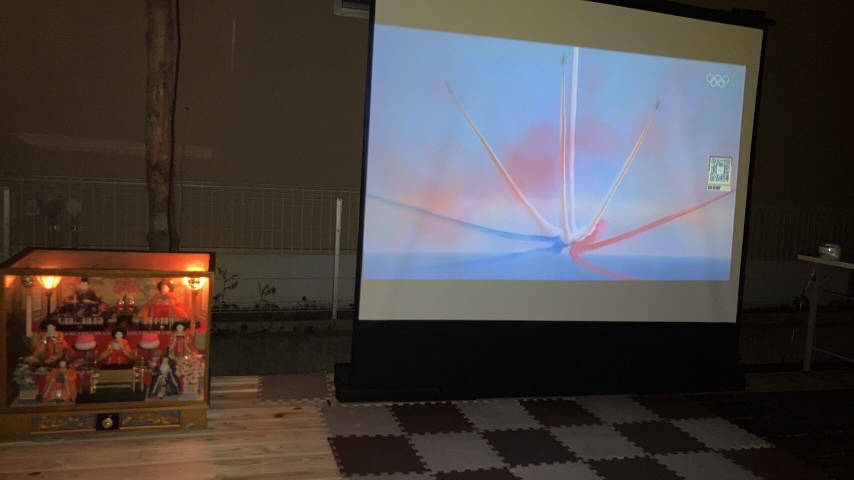 A projection screen displaying the Olympic rings logo, a QR code, and a blurred image of a sunset.