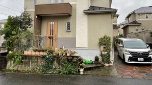 Un edificio residencial de dos pisos con entrada principal, un pequeño porche con plantas en macetas y un coche estacionado.