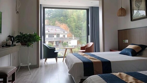 A hotel room with two beds, a desk, a chair, a potted plant, and a view of a building through a large window.