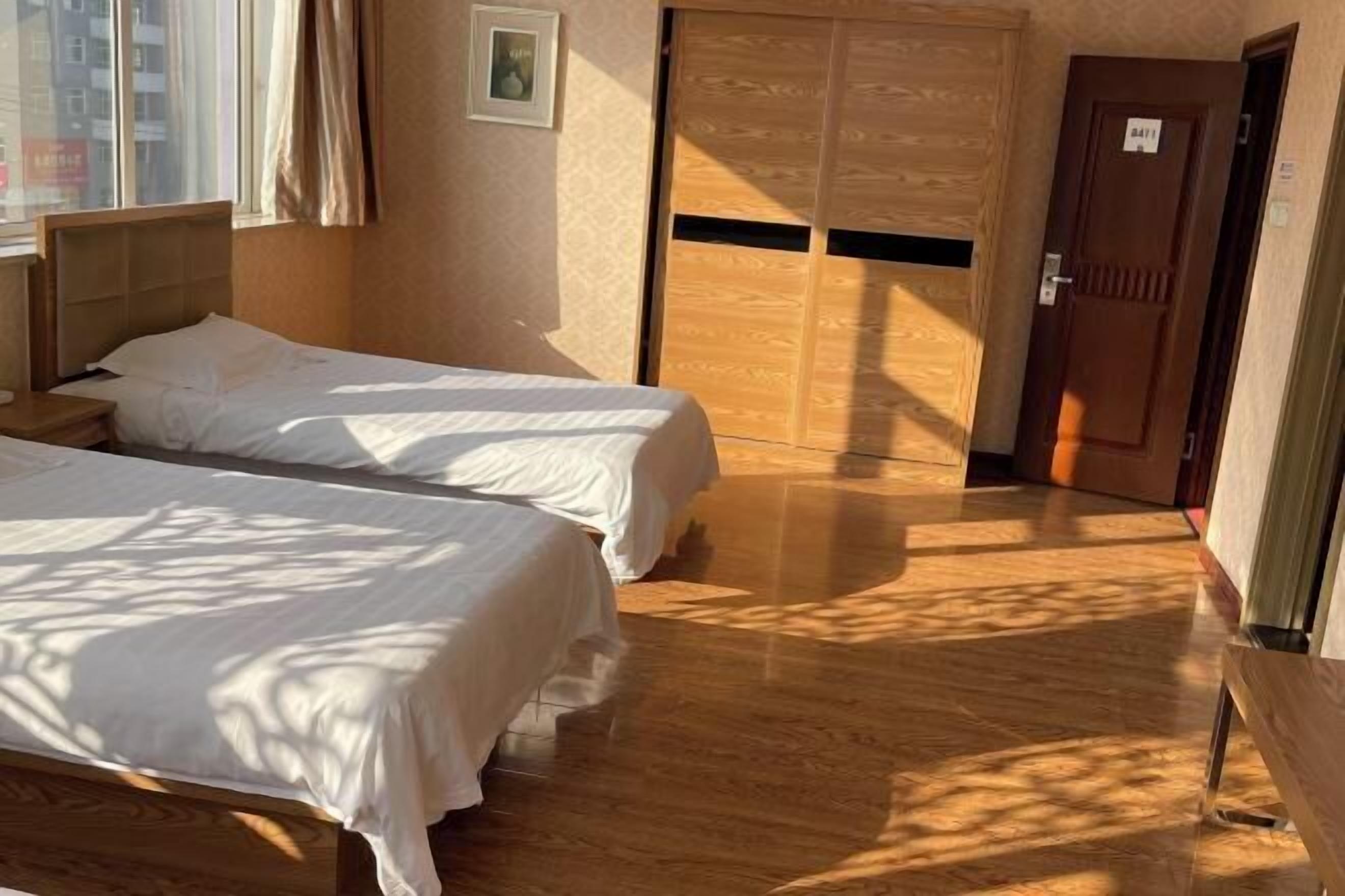 A hotel room with two beds, a wooden floor, a wardrobe, and a door with a sign.