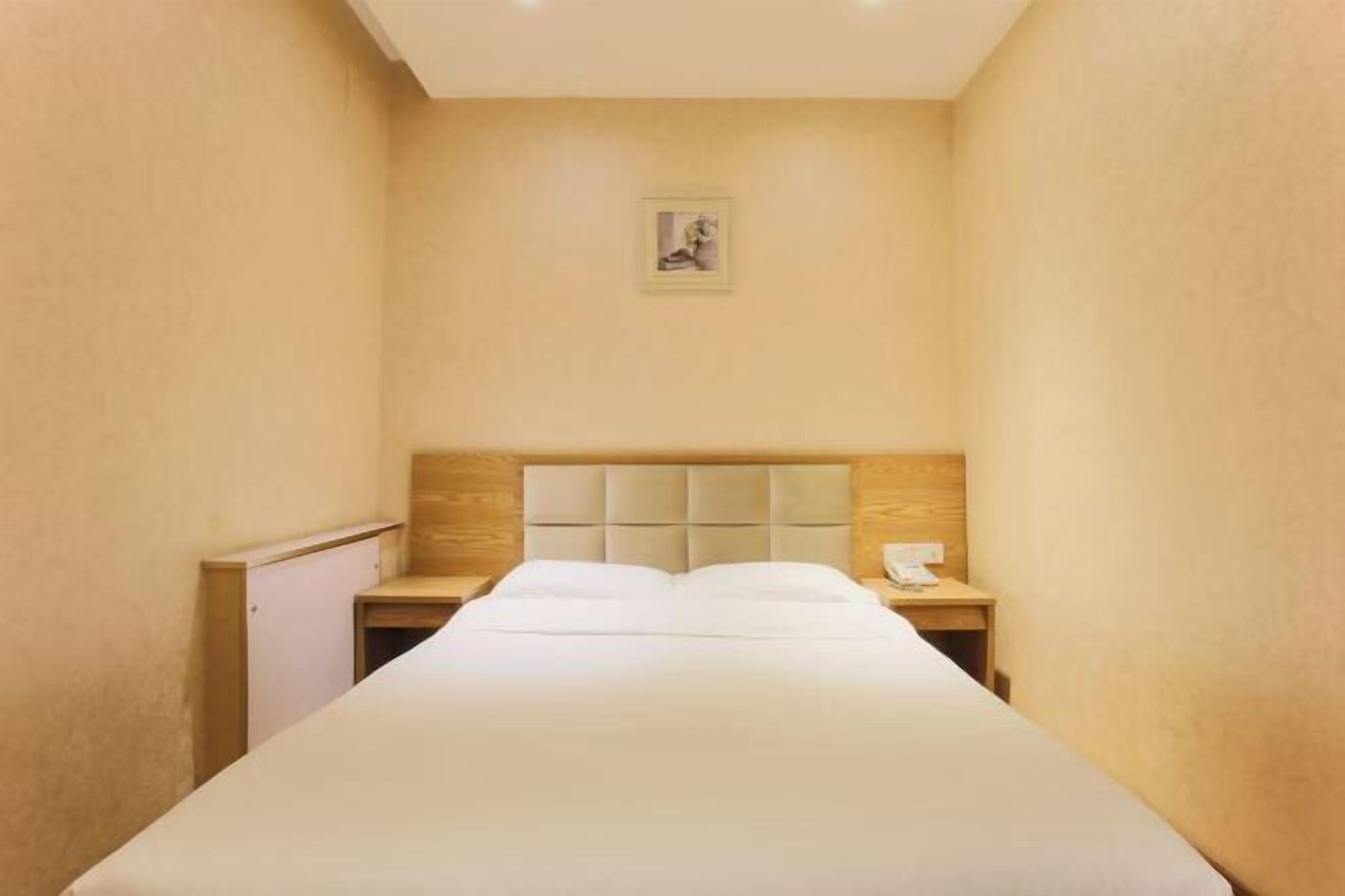 Une chambre d’hôtel étroite, avec un lit, une tête de lit en bois, une table de chevet et une petite table.