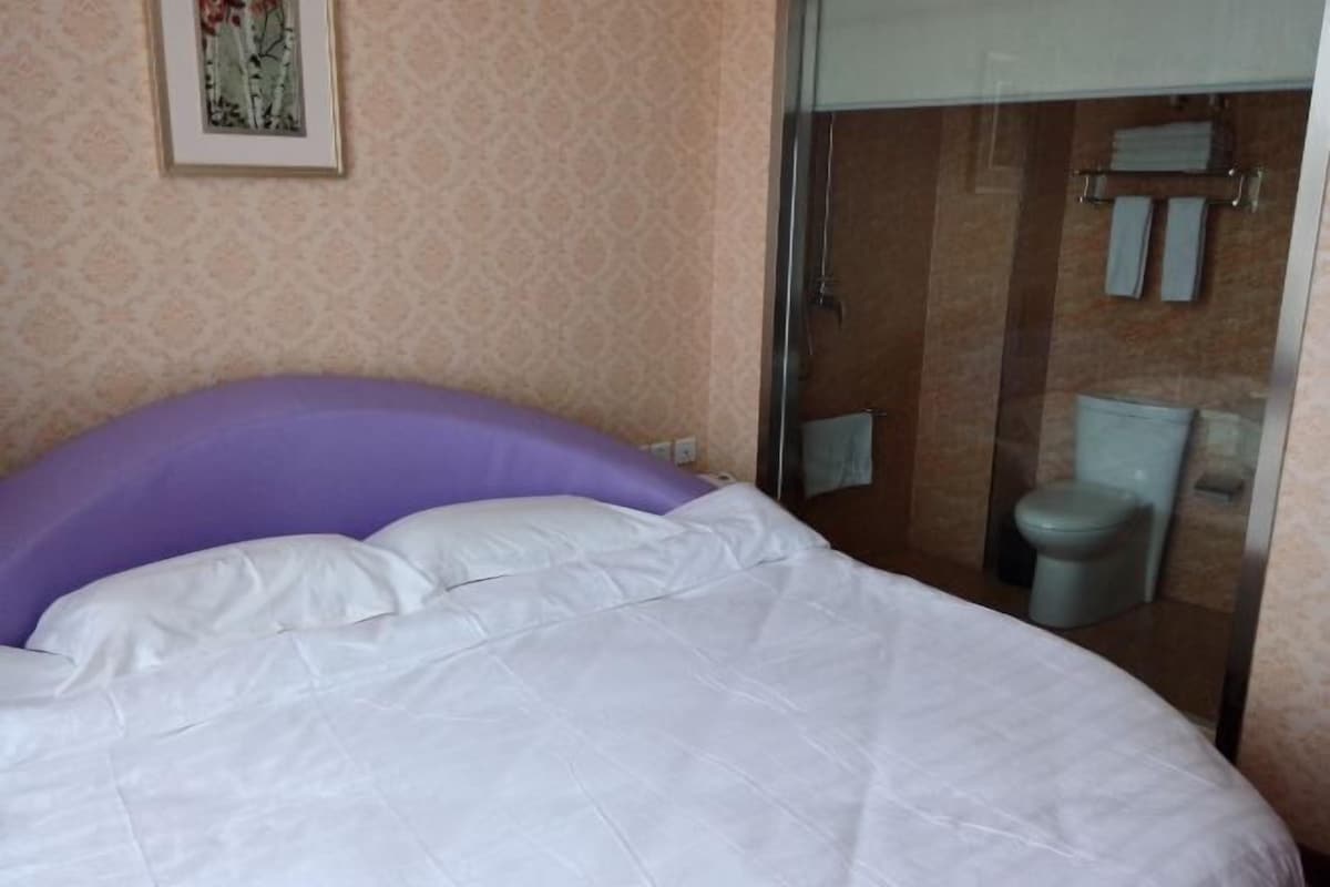 Un lit avec une tête de lit blanche et violette, une salle de bain avec une porte de douche en verre et des toilettes.