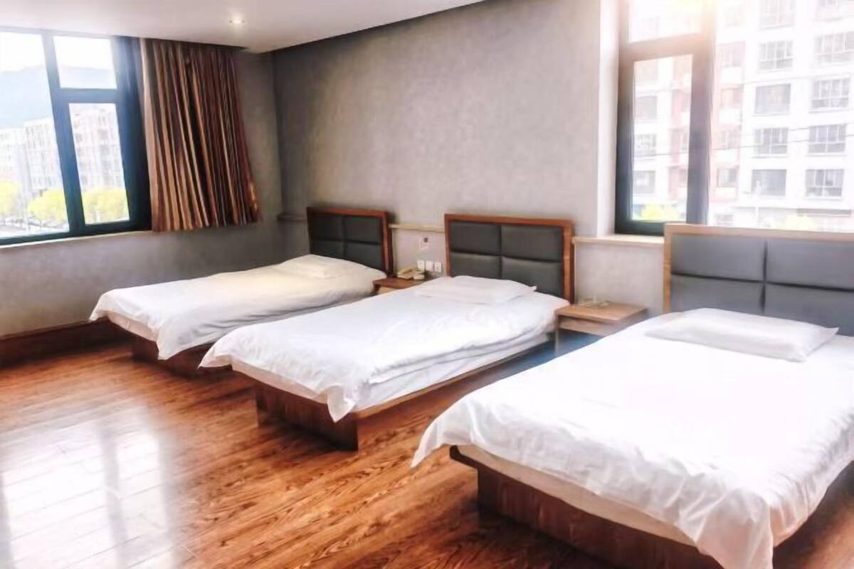 Ein Hotelzimmer mit drei Einzelbetten, Holzboden, einem großen Fenster mit Vorhängen und Blick auf die Stadt.
