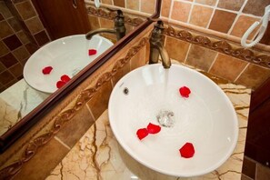 Un baño con lavamanos blanco, un espejo y pétalos de rosa sobre el suelo de mármol.