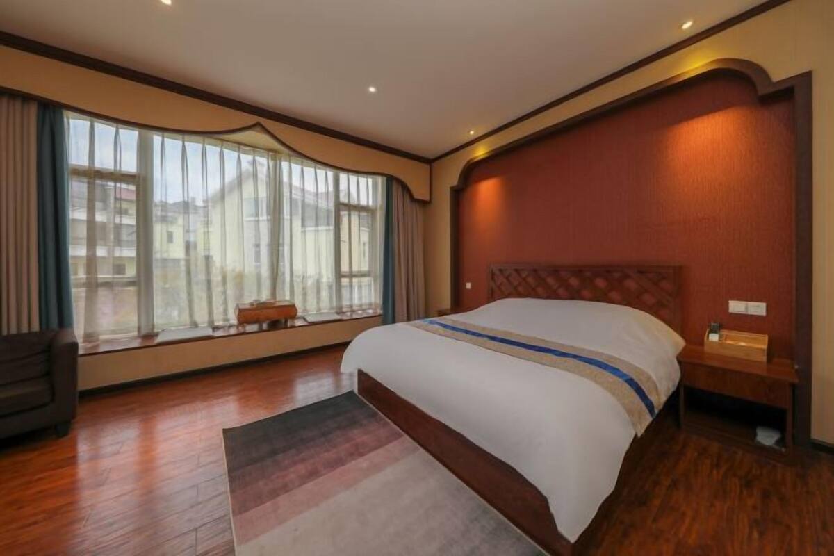 A hotel room with a large bed, a wooden floor, a window with curtains, and a nightstand with a lamp.
