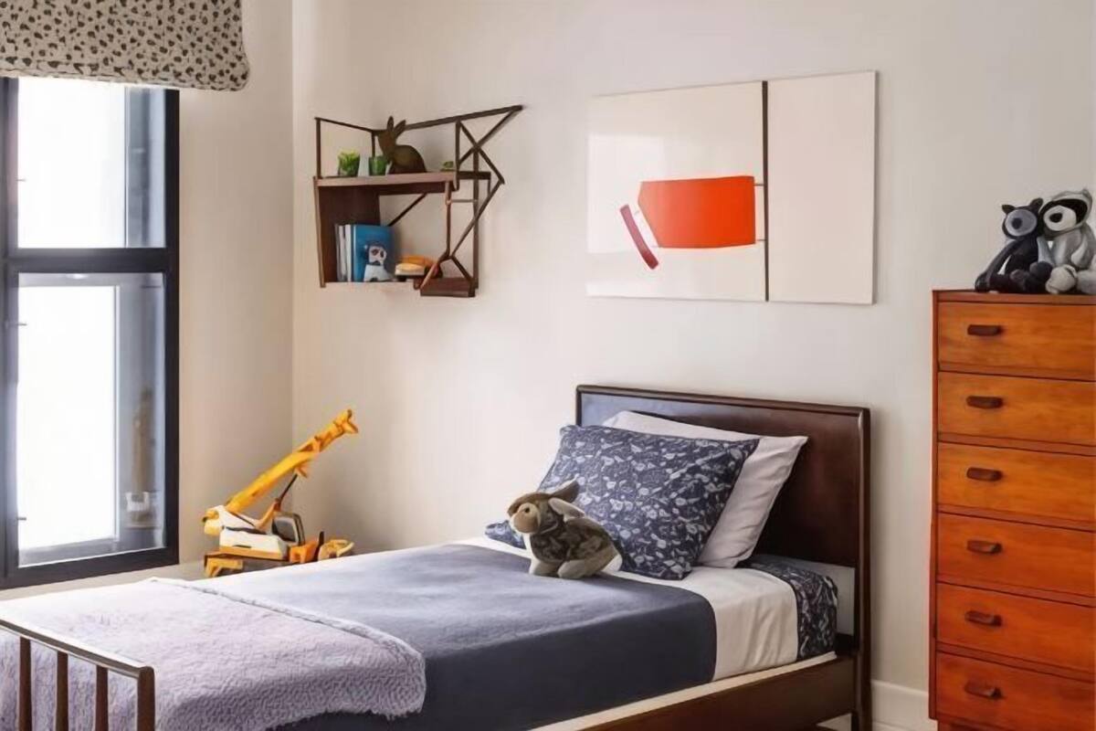 A bedroom with a bed, a wooden dresser, a shelf with books and toys, and a window with blinds.