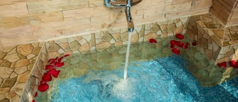 A stone-tiled bathtub with a faucet and rose petals floating on the water.