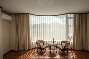 A room with a round table and two chairs, large windows with curtains, and an air conditioning unit on the wall.