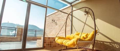 A room with a large glass door leading to a balcony, a hanging chair with yellow cushions, and a stone wall.